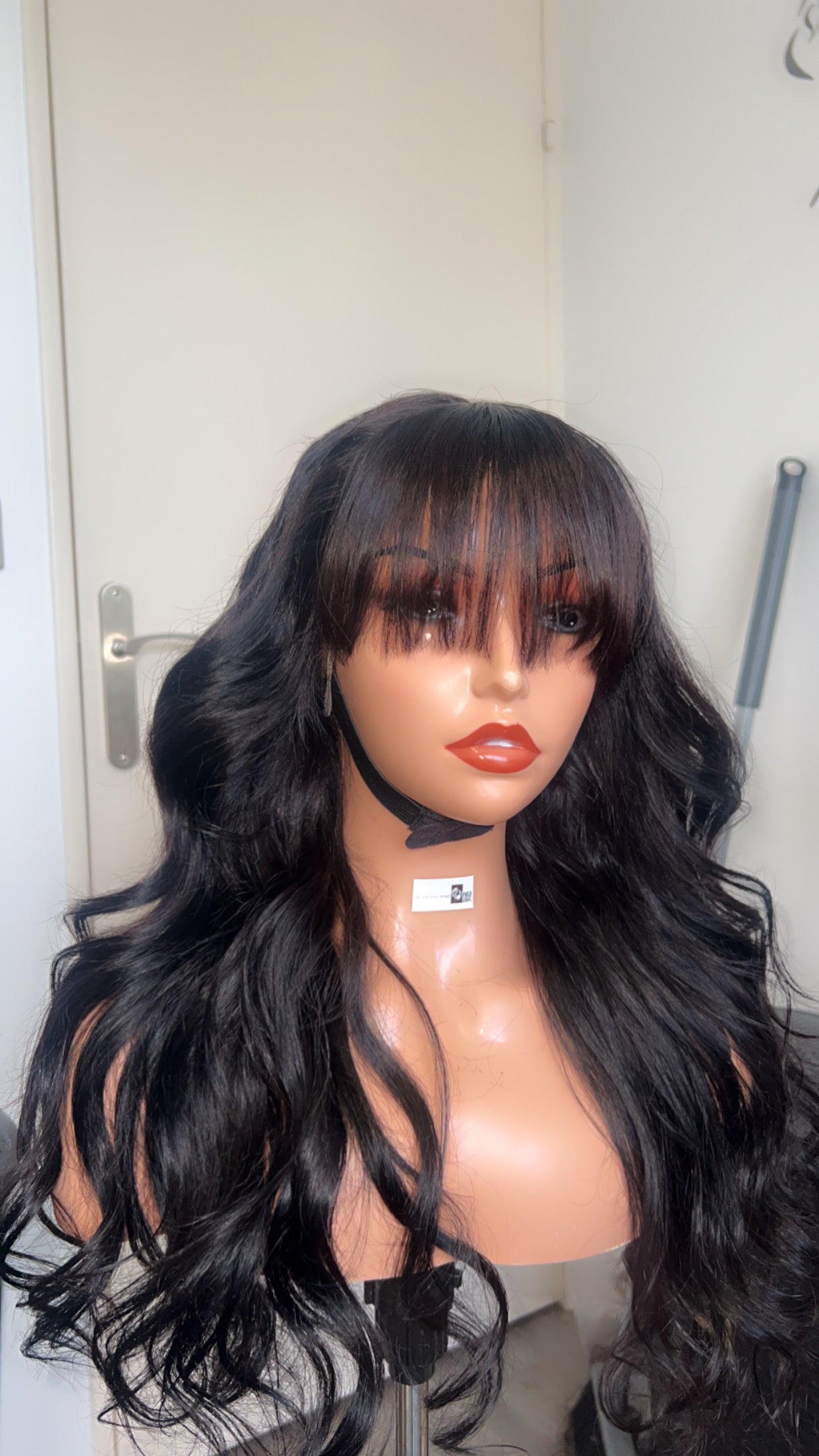Perruque ondulée avec frange - Mystère, avec des ondulations fluides et une frange moderne, en couleur noire intense, sur un mannequin. Produit haut de gamme pour un style chic et naturel.
